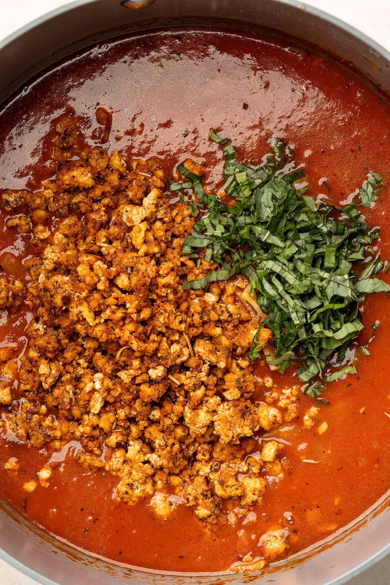 Tofu crumbles and chopped basil being added to large soup pot of lasagna soup.