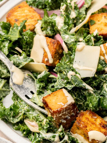Close up of a kale salad garnished with baked lemon herb tofu, pickled red onion, shaved parm on a serving plate with fork.