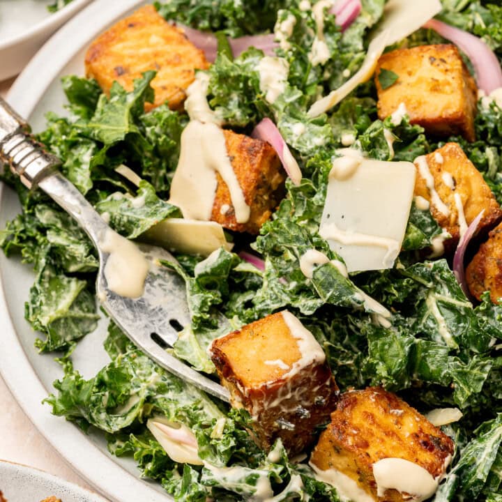 Close up of a kale salad garnished with baked lemon herb tofu, pickled red onion, shaved parm on a serving plate with fork.