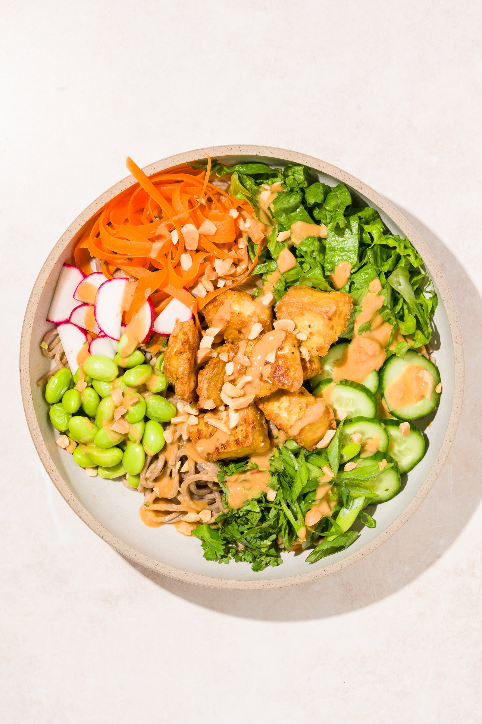 Bowl of soba noodles with crispy tofu and fresh vegetables and a drizzle of peanut sauce.