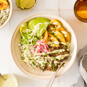 Close-up of caramelized plantains and black beans over rice with avocado, pickled red onions, corn, and jalapeño sauce.